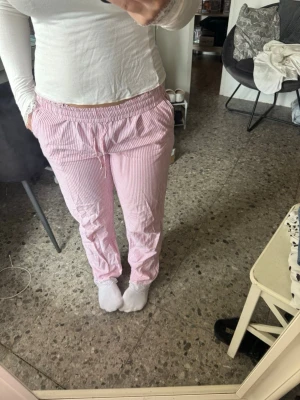Rosa- och vitrandiga pyjamasbyxor med spets  - Säljer ett par pyjamasbyxor i mjukt material med rosa och vita smala ränder och spets nedtill. Byxorna har resår och dragsko i midjan för extra komfort. Perfekta för mysiga hemmakvällar eller lata morgnar.