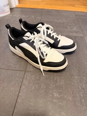 Svarta och vita Puma sneakers - Snygga sneakers från Puma i svart och vitt med klassisk låg siluett. Skorna har vita snören, svart sula och tydlig Puma-logga bak. Materialet är en mix av läder och textil för en sportig look. Aldrig använda. Kommer såklart med insulorna