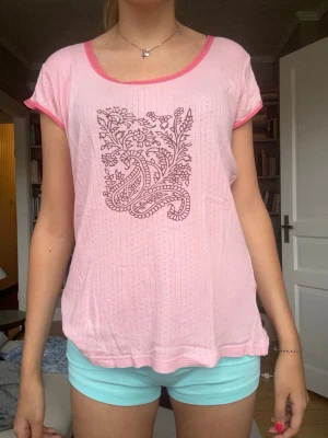 Ljusrosa topp med broderat mönster - Söt ljusrosa topp med korta ärmar och rund halsringning. Toppen har ett broderat blommönster framtill i mörkare nyans och kontrasterande rosa kant vid hals och ärmslut. Perfekt för varma dagar och enkel att styla till olika looks.