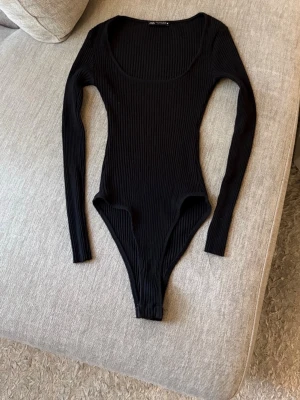 Svart ribbad body från Zara - Säljer en svart långärmad body från Zara i ribbat material. Bodyn har rundad urringning och är stretchig för en tight passform. Perfekt att styla med jeans eller kjol för en enkel men snygg look.