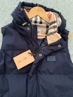 Mörkblå dunväst från Burberry - Snygg mörkblå dunväst från Burberry med klassiskt rutigt foder i beige, svart och rött. Västen har huva, dragkedja och tryckknappar samt en patch med Burberry-logga på bröstet. Perfekt för lager på lager och streetstyle.