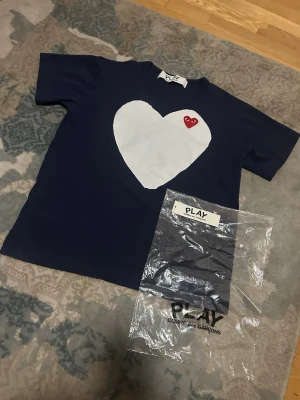 Blå Comme des Garçons PLAY t-shirt - Mörkblå t-shirt från Comme des Garçons PLAY med stort vitt hjärta och ett litet rött hjärta med ögon på bröstet. Klassisk rund hals och korta ärmar. Perfekt statement-plagg för dig som gillar streetwear och unika prints.
