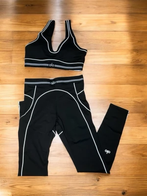 Svart träningsset från Alo - Snyggt svart träningsset från Alo med sport-bh och tights. Setet har vita kontrastsömmar som ger en sportig look. Tightsen har hög midja och fickor på sidorna. Materialet är stretchigt och passar perfekt för gym eller yoga.