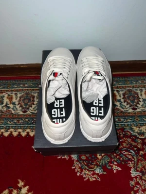 Vita sneakers från Tommy Hilfiger - Snygga vita sneakers från Tommy Hilfiger med klassisk låg profil. Skorna har vita snören, diskret logga på plösen och insida med svart, röd och vit detalj. Perfekta för en stilren och fräsch look. Materialet är slätt och lätt att hålla rent.