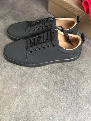 Svarta låga sneakers från Louboutin - Stilrena svarta sneakers från Christian Louboutin med mattsvart ovandel, svarta skosnören och beige innerfoder. Klassisk låg modell med diskret design och rundad tå. Perfekta för dig som gillar minimalistisk och clean stil.