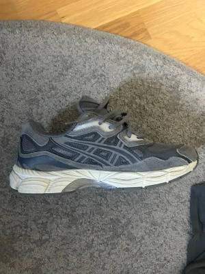 Grå Asics sneakers - Säljer ett par grå Asics sneakers med coola detaljer i mesh och mocka. Skorna har en chunky vit sula och klassiska Asics-loggor på sidan. Perfekta för dig som gillar sportig och trendig stil. Sköna att gå i och passar till det mesta.