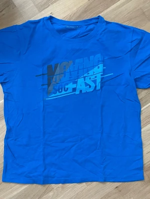 Blå t-shirt med tryck - Säljer en blå t-shirt med coolt tryck på bröstet där det står 'MOVING SO FAST' i olika blå nyanser. T-shirten har rund hals och korta ärmar. Perfekt för dig som gillar sportig och avslappnad stil.