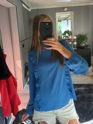 Blå blus med volangdetaljer - Supersnygg blå blus med långa ärmar och volangdetaljer längs framsidan. Perfekt att styla med jeans eller kjol! Den sitter som en S!