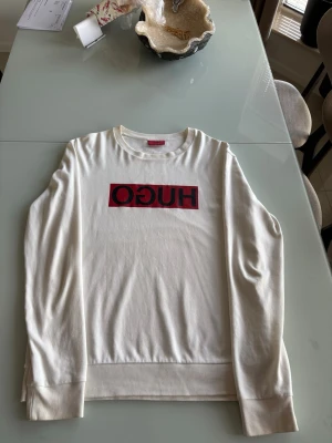 Vit sweatshirt med HUGO-logga - Snygg vit sweatshirt från HUGO med stor röd och svart logga på bröstet. Tröjan har rund halsringning, långa ärmar och ribbade muddar vid ärmslut och nederkant. Perfekt för en clean och modern look.