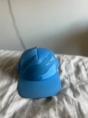 Blå Lacoste keps  - Snygg blå keps med stort krokodiltryck framtill. Kepsen har böjd skärm och mesh på sidorna för extra ventilation. Justerbar baktill för perfekt passform. Perfekt för dig som vill sticka ut med en cool accessoar.