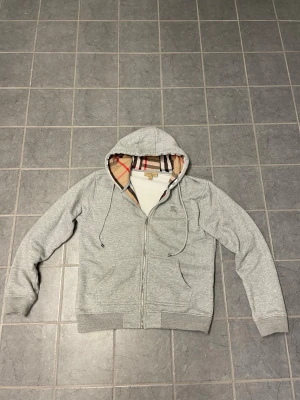 Grå hoodie från Burberry (passar som S/M) - Snygg grå zip-hoodie från Burberry med klassiskt rutigt foder i huvan och mjuk fleece på insidan. Hoodien har dragsko, två fickor fram och ribbade muddar. Perfekt för dig som vill ha en stilren och lyxig look.