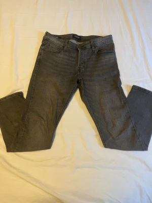 Grå slim glenn jeans Jack & Jones - Snygga grå slim fit jeans från Jack & Jones, modell SLIM/GLENN. Klassisk femficksdesign med dragkedjegylf och bälteshällor. Tillverkade i mjukt denimtyg som ger en bekväm passform och modern look.