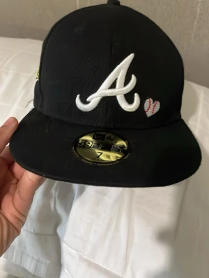 Svart Atlanta Braves keps 59FIFTY - Säljer en svart New Era 59FIFTY keps med Atlanta Braves logga i vitt framtill och ett litet hjärta med baseboll-sömmar. På sidan finns ett broderat 1995 World Series-märke i gult och vitt. Klassisk flat brim och broderade detaljer.