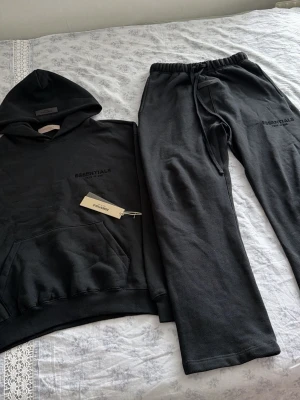Essentials tracksuit  - Säljer ett helt oanvänt svart set från Essentials med hoodie och matchande byxor, dustbag och tags medföljer. Hoodien har magficka och dragsko i huvan, byxorna har resår i midjan och dragsko. Båda delarna har Essentials-logga tryckt på bröst och lår. Mjukt och bekvämt material, perfekt för chill dagar. Byxorna och tröjan är i storlek S men som sitter oversized och därav passar även M. 