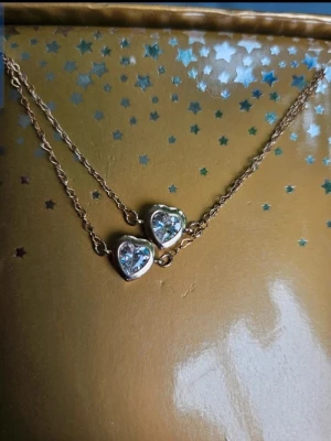 Guldigt halsband med hjärtan x2 - Säljer ett set med 2 likadana guldiga halsband med varsitt hjärtformat hänge med glittrande stenar. Kedjan är tunn och elegant, och halsbandet kommer i en snygg guldfärgad presentask med stjärnmönster. Perfekt accessoar för att lyfta vilken outfit som helst. Perfekt som vänskap, systrar eller mor och dotter  Kan även hämtas I Partille  J