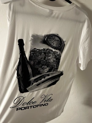 Vit Dolce Vita Portofino t-shirt - Snygg vit t-shirt med tryck av texten 'Dolce Vita Portofino' fram och ett stort svartvitt motiv på ryggen med flaska, stad och klocka. Klassisk rund hals och korta ärmar. Perfekt för dig som gillar stilrena plagg med coolt print.