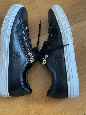 Rieker memsoft sneaker 41 - Stilrena svarta sneakers från Remonte med vita sulor, guldfärgade detaljer och dragkedja på sidan. Skorna har snörning och är tillverkade i skinnliknande material. Perfekta för dig som gillar en klassisk men trendig look.