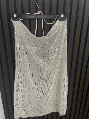 Glittrig silver kortklänning - Säljer en glittrig kortklänning i silver med paljetter. Klänningen har halterneck och ett snyggt fall framtill. Perfekt för dig som vill sticka ut och glänsa på nästa fest eller event.
