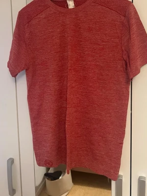 Röd träningst-shirt i funktionsmaterial - Säljer en röd kortärmad träningst-shirt i syntetmaterial. T-shirten har en melerad design och rund halsringning. Perfekt för gymmet eller löprundan.