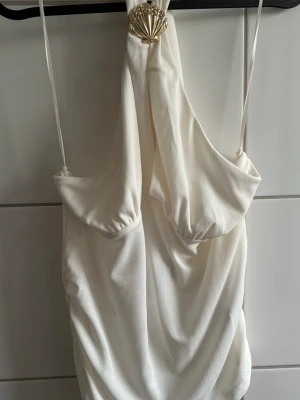 Vit top  med snäckdetalj - Snygg vit tip  med draperad design och tunna axelband. Framtill finns en guldfärgad snäckformad detalj som ger en exklusiv känsla. Materialet ser ut att vara mjukt och stretchigt, perfekt för strand eller poolhäng.