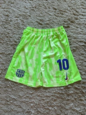 Neongröna FC Barcelona shorts - Säljer ett par neongröna FC Barcelona shorts med nummer 10 och Nike-logga framtill. Shortsen har elastisk midja och är tillverkade i ett lätt och ventilerande syntetmaterial, perfekta för fotboll eller träning.