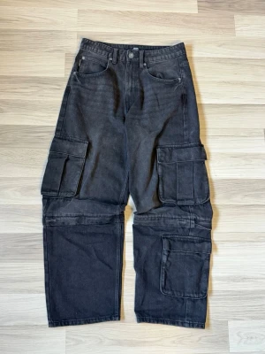 Svarta cargojeans - Säljer ett par svarta cargobyxor med bred passform och flera stora fickor på sidorna. Byxorna har raka ben, bälteshällor och klassisk jeansknäppning. Materialet är kraftigt och slitstarkt, perfekt för en streetwear-look.