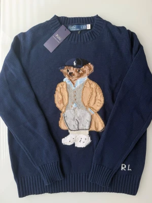 Mörkblå stickad tröja Polo Bear (oldmoney) - Mörkblå stickad tröja från Polo Ralph Lauren med den ikoniska Polo Bear-björnen broderad framtill. Tröjan har rund hals, ribbade muddar och RL-broderi vid ärmslutet. Perfekt för dig som gillar klassisk och lekfull stil.
