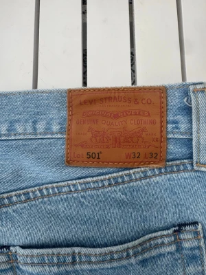 Levi's 501 ljusblå raka jeans - Klassiska Levi's 501 jeans i ljusblå denim med rak passform och knappgylf. Jeansen har fem fickor och den ikoniska läderpatchen bak i midjan. Perfekta för en avslappnad och tidlös stil.