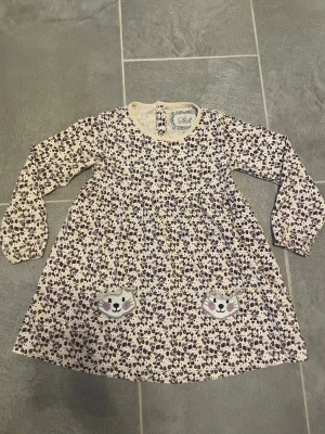 Blommig klänning med kattfickor Skill - Söt blommig klänning från Skill i vitt och lila med lång ärm och två fickor framtill med kattmotiv. Perfekt för dig som gillar gulliga detaljer och lekfull stil. Klänningen har rund hals och är gjord i mjukt material.