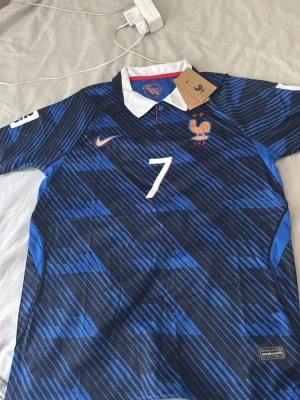 Frankrike fotbollströja Dembele #7 - Snygg blå fotbollströja från Nike med Frankrikes landslagsmärke och nummer 7, Dembele på ryggen. Tröjan har vit krage, orange Nike-logga och ett mönster i olika blå nyanser. Perfekt för match eller träning.
