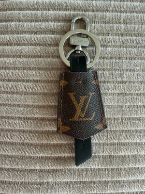 Vuitton nyckelring brun monogram - Säljer en exklusiv nyckelring från  Vuitton i klassiskt brunt monogrammönster med LV-logga i metall. Nyckelringen har detaljer i silverfärgad metall och svart läder. Perfekt accessoar för att lyfta din väska eller nyckelknippa.