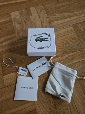 Silverfärgat armband från Lacoste - Snyggt och stilrent armband i silverfärg från Lacoste med den ikoniska krokodilen som hänge. Kedjan är tunn och justerbar, vilket gör att den passar de flesta handleder. Kommer med original ask, påse och tags.