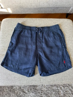 Mörkblå linneshorts från Ralph Lauren  - Snygga mörkblå shorts i linne med elastisk midja och snörning framtill. Klassisk passform och diskret röd logga broderad på benet. Perfekta för varma dagar och enkel att matcha med olika stilar. SMÅ I STORLEK!