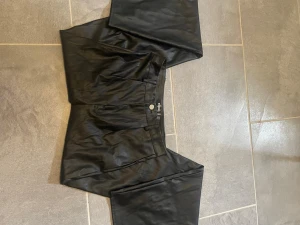 Svarta byxor från Missguided - Säljer ett par svarta byxor från Missguided med hög midja och raka ben. Byxorna har en snygg glansig finish som ger en cool look och stängs med knapp och dragkedja. Perfekta för dig som vill sticka ut lite extra.
