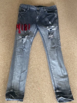 Amiri jeans - Säljer ett par grå slim fit jeans med slitningar och hål på framsidan. På vänster lår finns röd broderad text 'MIRI!' och på baksidan en röd broderad detalj på fickan. Jeansen har klassisk femficksmodell och är i tvättad denim.