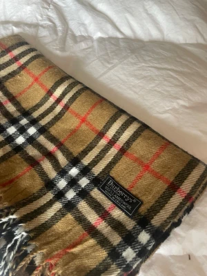 Klassisk beige rutig halsduk Burberry - Säljer en ikonisk beige halsduk från Burberry med det klassiska rutmönstret i svart, vitt och rött. Halsduken har fransar i ändarna och är tillverkad i mjuk ull. Perfekt accessoar för att lyfta vilken outfit som helst.