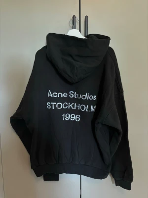 Svart hoodie Acne Studios Stockholm - Säljer en svart hoodie från Acne Studios med stor huva och texten 'Acne Studios STOCKHOLM 1996' tryckt på ryggen. Hoodien har avslappnad passform, ribbade muddar och är tillverkad i mjukt bomullsmaterial. Perfekt för en cool och enkel streetwear-look.