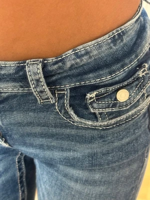 Blå jeans med broderade detaljer - Snygga blå jeans med låg midja och unika broderade detaljer samt paljetter på bakfickan. Jeansen har klassisk femficksdesign och kontrastsömmar. Perfekta för dig som vill sticka ut med coola detaljer och en trendig look.
