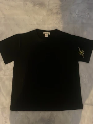 Svart t-shirt med liten logga - Snygg svart oversized t-shirt med korta ärmar och rund hals. På vänster ärm finns en liten gul logga som ger en cool detalj. Perfekt till jeans eller shorts för en clean och stilren look.