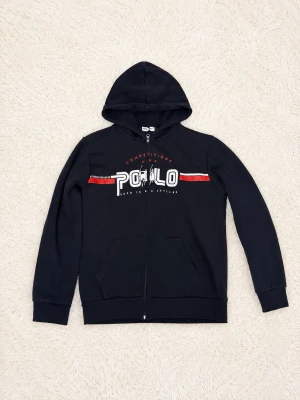 Svart hoodie polo hoodie storlek L - Snygg svart hoodie med dragkedja och huva. På bröstet finns ett stort vitt och rött POLO-tryck samt texten 'COMPETITIONS'. Hoodien har två fickor framtill och ribbade muddar vid ärmar och nederkant. Perfekt för en avslappnad streetstyle-look.