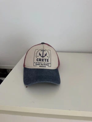 Beige och vinröd keps - Säljer en keps med beige frontpanel och mörkblå skärm. Sidorna och bakstycket är vinröda. Framtill finns ett tryck med texten 'CRETE' och ett ankare. Perfekt för dig som gillar en avslappnad stil. Köpt i kreta (Grekland)