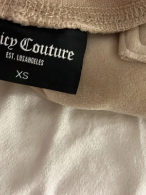 Beige sett från Juicy Couture - Säljer ett beige sett från Juicy Couture i storlek XS. Setet består av en zip-hoodie med fickor och matchande byxor och fickor. Båda delarna har broderad logga. Perfekt för chill dagar hemma eller när du vill vara extra comfy.