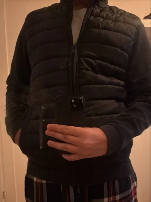 Svart dunjacka med quiltade paneler - Säljer en svart dunjacka med quiltade paneler och dragkedja framtill. Jackan har hög krage, två sidofickor och långa ärmar i mjukt material. Perfekt för kalla dagar och ger en clean och stilren look.