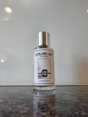 LEBLANC 24.0 Eau de Parfum - Minimalistisk parfymflaska i genomskinligt glas med metallock. Doften heter LEBLANC 24.0 och etiketten har en stilren design med svart text och grafiskt tryck. Flaskan har en modern och enkel form som passar perfekt på badrumshyllan.