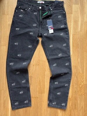 Svarta hilfiger jeans med broderade loggor - Säljer ett helt nytt par svarta jeans från Tommy Jeans med vita broderade loggor över hela byxan. Klassisk femficksmodell med raka ben och knappgylf. 