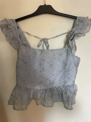 Ljusblå blommig blus med volang - Supersöt ljusblå blus med blommigt mönster, volangkant nertill och volangärmar. Toppen har knytning i ryggen och är tillverkad i ett tunt, lätt material som känns luftigt och somrigt.