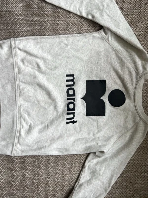 Grå hoodie med svart Marant-tryck - Snygg grå hoodie med stort svart Marant-tryck på bröstet. Hoodien har en klassisk passform med långa ärmar och ribbade muddar. Perfekt för en avslappnad och trendig look. Materialet är mjukt och bekvämt, troligen bomullsblandning.