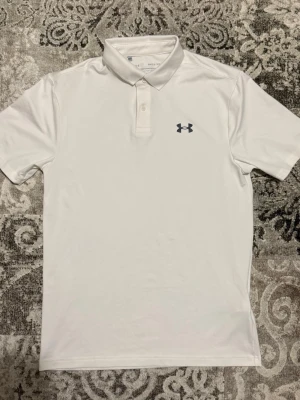 Vit performance piké från Under Armour - Säljer en vit performance pikétröja från Under Armour med klassisk krage och knappar framtill. Tröjan har korta ärmar och är tillverkad i ett lätt och funktionellt material som passar perfekt för sportiga aktiviteter.