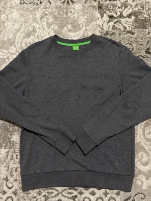 Mörkgrå sweatshirt från Hugo Boss - Mörkgrå sweatshirt från Hugo Boss med diskret broderad logga på bröstet. Tröjan har rund halsringning, långa ärmar och ribbade muddar vid ärmslut och nederkant. Insidan av kragen har en grön detalj. Perfekt för en stilren och avslappnad look.