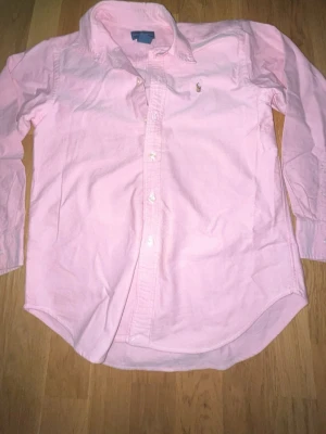Ljusrosa skjorta från Ralph Lauren - Säljer en klassisk ljusrosa skjorta från Ralph Lauren med broderad logga på bröstet. Skjortan har lång ärm, knappar framtill och en tidlös krage. Perfekt för dig som vill ha en stilren och fräsch look.
